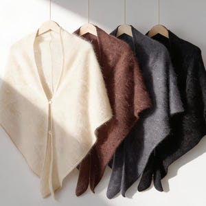 Może przedstawiać: Cztery miękkie, puszyste szale w kolorze kremowym, brązowym, szarym i czarnym wiszą na drewnianych wieszakach. Każdy szal ma zapięcie na guziki przy dekolcie. Szale wykonane są z teksturowanego materiału, prawdopodobnie wełny lub moheru.