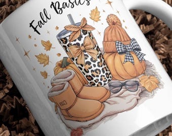 May include: White ceramic mug with the text "Fall Basics" and autumn-themed illustrations. The design includes a leopard print tumbler, a pumpkin, boots, a hat, and sunglasses. The mug is presented in a white box with brown paper filler.