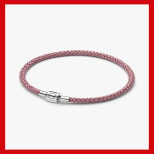 Puede incluir: Una pulsera trenzada de color rosa empolvado con un cierre cilíndrico de metal plateado. El cierre lleva grabado el texto PANDORA. La pulsera tiene forma circular y una estructura de cordón tejido con textura en toda la banda.