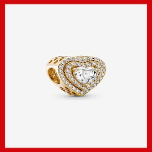 May include: A gold-colored heart-shaped charm with a clear, heart-shaped stone in the center. The charm is surrounded by a double halo of small, clear stones. The sides of the charm have a decorative openwork design.