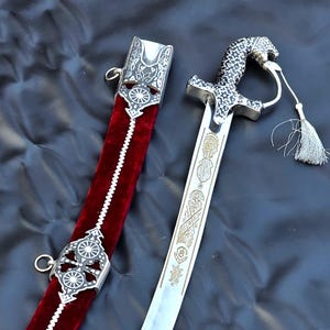 May include: A curved steel sword with an ornate silver-toned hilt shaped like a feline head and a matching scabbard covered in deep red velvet. The scabbard features intricate silver metal fittings with circular patterns and a white tassel.