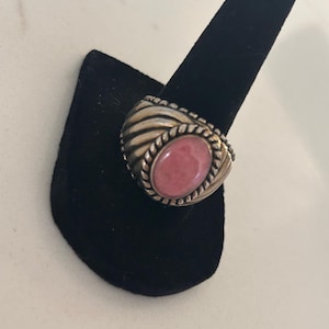 May include: Silver ring featuring a large, oval pink gemstone. The ring has a textured band with a braided border around the stone. The ring is displayed on a black velvet ring holder.