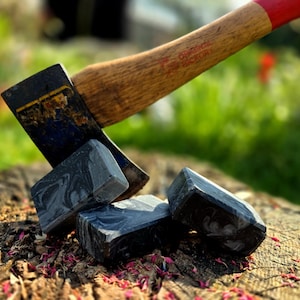 Könnte beinhalten: Eine Axt mit Holzgriff und rotem Griff ist in einen Baumstumpf eingeschlagen. Mehrere dunkelgraue, rechteckige Seifenstücke mit Marmormuster liegen um die Axt herum.