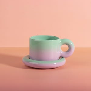 May include: A two-tone ceramic mug and saucer set. The mug has a distinctive rounded handle and a colour gradient from mint green to pink. The saucer matches the mug's colour scheme. The set is displayed on a pink surface.
