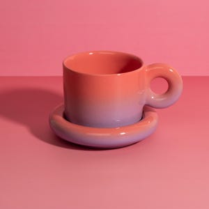 Può includere: Un set di tazza e piattino da caffè in ceramica con un'esclusiva maniglia a forma di ciambella e un piattino abbinato. La tazza e il piattino presentano una sfumatura di colore, che va dal corallo al lavanda. Il set è posizionato su una superficie rosa.