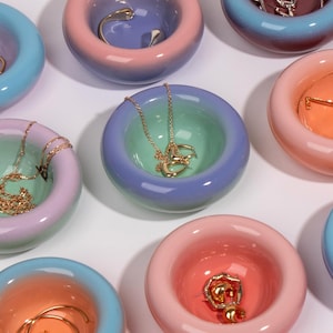 Op de afbeelding: Een verzameling kleurrijke, ronde keramische sieradenbakjes. Elk bakje heeft een kleurovergang van blauw, roze en groen. Verschillende gouden kettingen en ringen worden in de bakjes getoond, wat hun gebruik als decoratieve opslag aantoont.