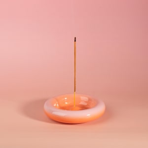 May include: A ceramic incense holder, donut-shaped, with an orange to white gradient. An incense stick stands upright in the centre, set against a soft pink backdrop. The holder is a decorative item for aromatherapy, measuring approximately 10 cm in diameter.
