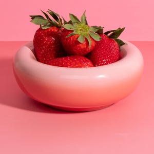 Op de afbeelding: Een roze keramische kom gevuld met verse, rijpe aardbeien. De kom heeft een afgeronde vorm met een kleurverloop, variërend van lichtroze tot een dieper rood. De aardbeien zijn helderrood met groene bladeren.