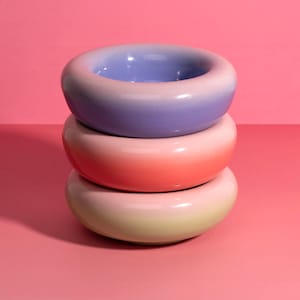 Könnte beinhalten: Drei gestapelte, donut-förmige Keramikschalen mit Farbverlauf. Die obere Schale ist lila, die mittlere korallenfarben und die untere hellgelb. Die Schalen stehen auf einer rosa Oberfläche vor einem rosa Hintergrund.