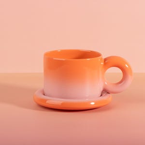 May include: A ceramic mug and saucer set with a gradient color scheme. The mug transitions from orange to pink, featuring a unique circular handle. The saucer mirrors the mug's color palette, creating a cohesive design.