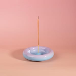 Op de afbeelding: Een keramische wierookhouder in een blauw-paarse gradiënt, met een brandende wierookstok. De houder heeft een afgeronde, donut-achtige vorm. De wierookstok is bruin met een gele basis. De achtergrond is effen perzikkleurig.