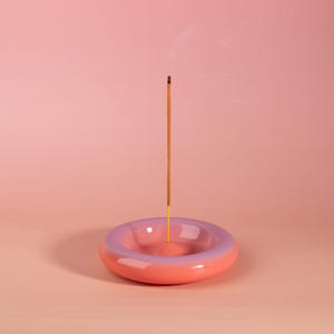May include: A ceramic incense holder in a donut shape, with a gradient of pink and purple. An incense stick is lit and emitting smoke. The background is a solid pink.