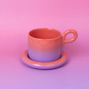Peut inclure: Un ensemble tasse et soucoupe en céramique avec un dégradé de couleurs. La tasse passe d'un rose corail en haut à un violet lavande à la base. La soucoupe est violette lavande assortie. La anse est une boucle arrondie.