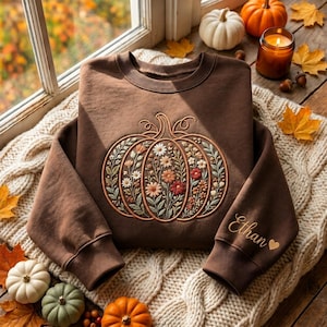Pode incluir: Uma camisola castanha de manga comprida com um grande desenho de abóbora bordado e preenchido com várias flores na parte da frente. O nome Ethan e um pequeno símbolo de coração estão bordados em letra cursiva no punho da manga esquerda.