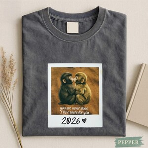 Puede incluir: Camiseta gris oscuro con una imagen impresa estilo polaroid de dos monos besándose. La imagen incluye el texto "you are never alone, I'll be there for you" y "2026". La camiseta está doblada cuidadosamente.