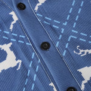 K&ouml;nnte beinhalten: Eine blaue Strickjacke mit Knopfleiste, verziert mit einem wiederkehrenden wei&szlig;en Hirschmuster und hellblauen, gestrichelten Rautenlinien. Das Kleidungsst&uuml;ck hat eine gerippte Textur und drei sichtbare schwarze Kn&ouml;pfe an der Knopfleiste.