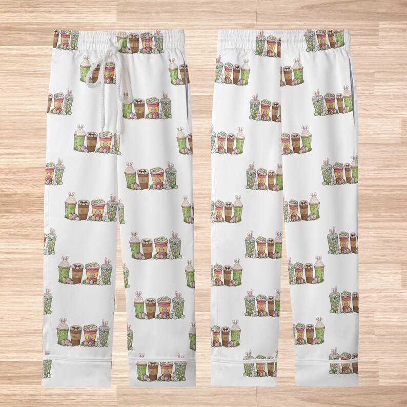 May include: White pajama pants with a repeating pattern of illustrated coffee cups, some with bunny ears, in shades of brown, green, and pink. The pants have a drawstring waist and a relaxed fit.