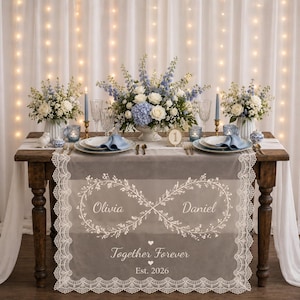 May include: A gray table runner with white lace trim featuring an embroidered infinity symbol made of leaves, the names Olivia and Daniel, the text Together Forever, and Est. 2026. The runner is displayed on a wooden table with floral centerpieces.