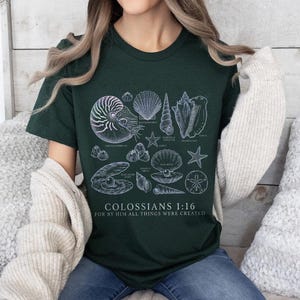 May include: Dark green t-shirt featuring a detailed illustration of various seashells in white. The design includes a nautilus, starfish, and other marine life. The text "COLOSSIANS 1:16 FOR BY HIM ALL THINGS WERE CREATED" is printed below the illustrations.