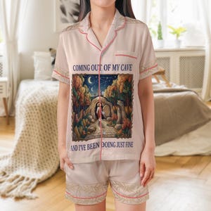 Può includere: Pigiama beige con maniche corte e pantaloncini. Il set presenta finiture rosa e una grafica sul davanti con la scritta "COMING OUT OF MY CAVE" e "AND I'VE BEEN DOING JUST FINE". La grafica raffigura una persona che esce da una grotta.