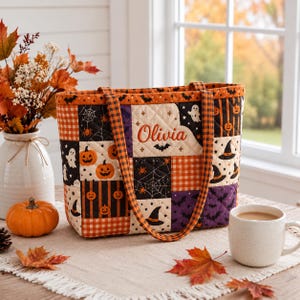 May include: A quilted tote bag featuring a patchwork Halloween pattern with pumpkins, ghosts, bats, spiders, and webs. The bag has orange and black plaid straps and the name Olivia embroidered in orange thread on a cream-colored fabric panel.