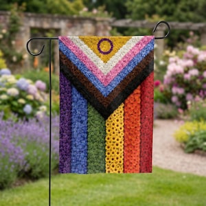 May include: A rectangular garden flag featuring a vertical rainbow stripe pattern overlaid with a chevron design composed of small flowers in shades of yellow, white, pink, light blue, brown, and black, with a purple circle in the center.