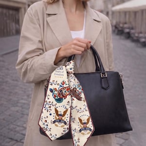 May include: A black leather handbag with a decorative scarf featuring a patriotic design. The scarf displays an eagle, floral patterns, and the text "250 Years of Freedom" and "1776-2026". The woman is wearing a tan trench coat.