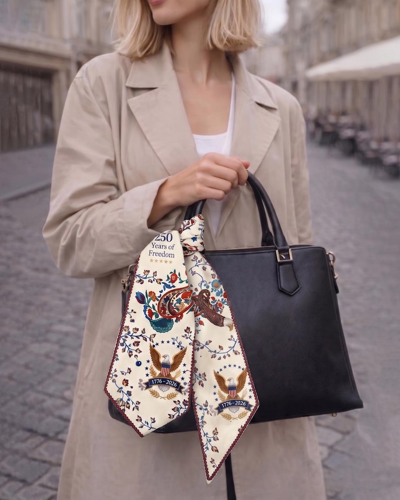 May include: A black leather handbag with a decorative scarf featuring a patriotic design. The scarf displays an eagle, floral patterns, and the text "250 Years of Freedom" and "1776-2026". The woman is wearing a tan trench coat.