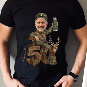 Puede incluir: Camiseta negra con una ilustración de dibujos animados de un hombre con ropa de camuflaje sosteniendo un rifle y un termo con el nombre "Liam". El hombre está sentado en un gran "50" con un ciervo y una mochila. También está el texto "50 YEARS".
