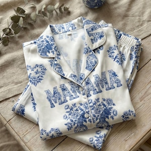 Może przedstawiać: Biały zestaw piżamowy z niebieskim wzorem kwiatowym i wazonem. Słowo "MAMA" jest wydrukowane dużymi niebieskimi literami z przodu góry. Zestaw zawiera koszulę zapinaną na guziki i pasujące spodnie.