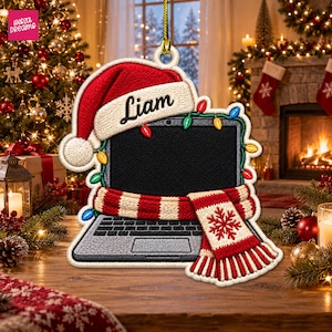 May include: A flat, ornament-style laptop decoration featuring a red Santa hat with the name Liam embroidered on the brim. The laptop is wrapped in a string of colorful Christmas lights and a red and white striped scarf with a snowflake pattern.
