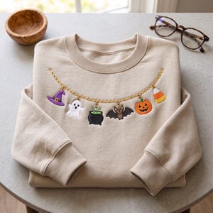 May include: A beige crewneck sweatshirt featuring an embroidered gold chain graphic across the chest. The chain displays dangling charms including a purple witch hat, a white ghost, a black cauldron, a brown bat, an orange pumpkin, and candy corn.