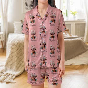 May include: Pink pajama set featuring a repeating pattern of a brown cow wearing bunny ears and sunglasses, with the name "Sophia" in cursive script. The set includes a button-down shirt and shorts.