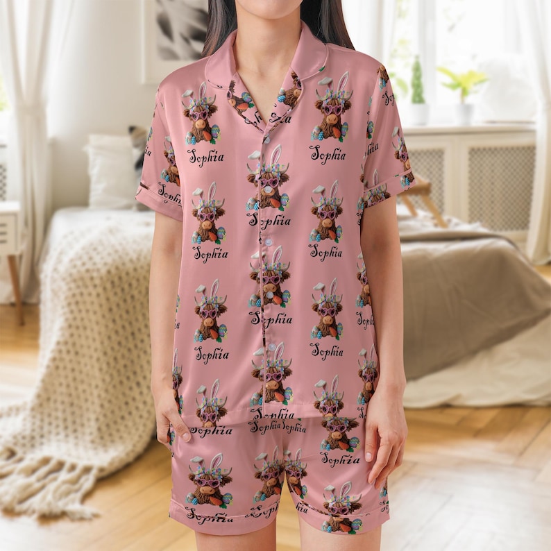 May include: Pink pajama set featuring a repeating pattern of a brown cow wearing bunny ears and sunglasses, with the name "Sophia" in cursive script. The set includes a button-down shirt and shorts.