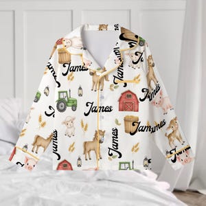 Puede incluir: Pijama blanco con un estampado de animales de granja, incluyendo caballos, cerdos, ovejas y un tractor. La palabra "James" está escrita en cursiva sobre la tela. El pijama tiene cuello y ribetes amarillos.