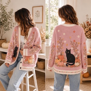 K&ouml;nnte beinhalten: Eine lang&auml;rmelige, gekn&ouml;pfte Strickjacke in einem zarten Rosaton. Das Kleidungsst&uuml;ck ist mit einer Stickerei versehen, die eine schwarze Katze inmitten von orangefarbenen K&uuml;rbissen und verschiedenen Wildblumen zeigt. Die Jacke hat einen cremefarbenen Saum.
