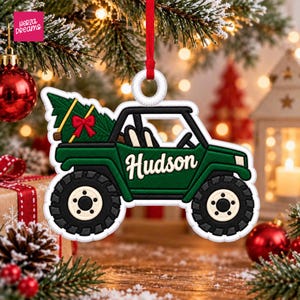 May include: A green off-road vehicle Christmas ornament featuring a dark green body, large black tires, and a green pine tree in the back with a red bow. The name Hudson is printed in white script on the side of the vehicle. It hangs from a red ribbon.