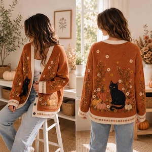 K&ouml;nnte beinhalten: Eine rostorange Strickjacke mit Stickereien von einer schwarzen Katze, K&uuml;rbissen und Blumen. Das Kleidungsst&uuml;ck hat einen V-Ausschnitt, lange &Auml;rmel und eine Knopfleiste mit cremefarbenen Abschl&uuml;ssen an den Kanten und &Auml;rmelb&uuml;ndchen, getragen zu Jeans.
