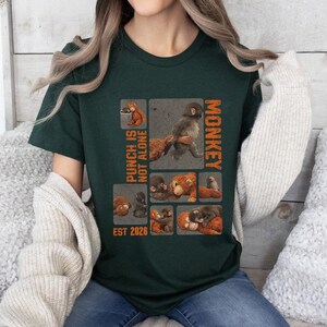 Könnte beinhalten: Dunkelgrünes T-Shirt mit einem Affen-Motiv und dem orangefarbenen Text "PUNCH IS NOT ALONE MONKEY". Das Design umfasst Affenillustrationen und den Text "EST 2026". Das T-Shirt besteht aus einem weichen Material.