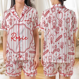 Könnte beinhalten: Weiß-rotes gestreiftes Pyjama-Set mit einem Schulmotiv. Das Set besteht aus einem kurzärmeligen Hemd mit Knöpfen und Shorts, beide rot eingefasst. Das Hemd trägt den Namen "Rosie" in roter Schrift.
