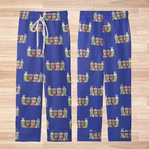 May include: Blue pajama pants with a repeating pattern of various coffee drinks. The drinks are in shades of brown, green, and white. The pants have a drawstring waist and are displayed on a light brown wooden surface.