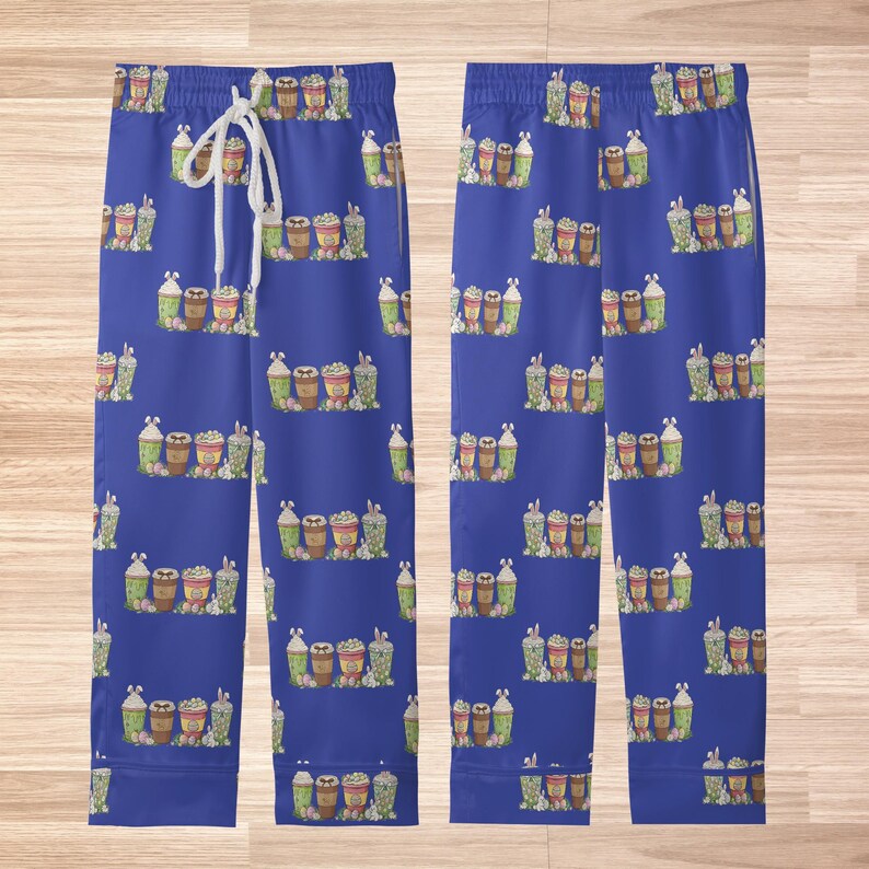 May include: Blue pajama pants with a repeating pattern of various coffee drinks. The drinks are in shades of brown, green, and white. The pants have a drawstring waist and are displayed on a light brown wooden surface.