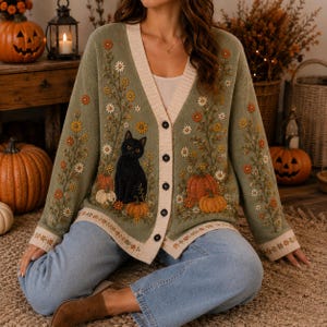 K&ouml;nnte beinhalten: Eine olivgr&uuml;ne Strickjacke mit V-Ausschnitt, Knopfleiste und cremefarbenem Saum. Das Strickkleidungsst&uuml;ck ist mit gestickten Blumenranken, orangefarbenen K&uuml;rbissen und einer schwarzen Katze auf den Vorderteilen und den langen &Auml;rmeln verziert.