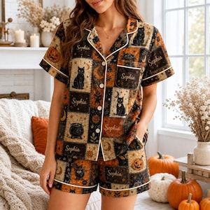May include: A short-sleeved button-down pajama set featuring a patchwork pattern of black cats, pumpkins, cauldrons, and crescent moons in orange, black, and cream. The name Sophia is printed in script across several of the patterned squares.