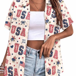 Puede incluir: Camisa crema con botones con un tema patriótico de EE. UU. La camisa presenta letras a cuadros rojas y azules que deletrean "USA", junto con patrones florales y de estrellas, banderas estadounidenses y el texto "EST. 1776".