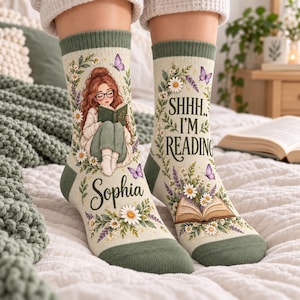 May include: Cream-colored crew socks with a green ribbed cuff and toe. One sock features an illustration of a person with red hair reading a book, the name Sophia, and floral accents. The other sock displays the text SHHH.. I'M READING with an open book and flowers.
