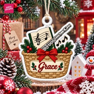 Puede incluir: Un adorno navideño colgante con forma de cesta tejida que presenta una flauta plateada, un libro de partituras con clave de sol, ramas de pino, bayas rojas y un lazo rojo. La cesta tiene el nombre Grace bordado en letra roja.