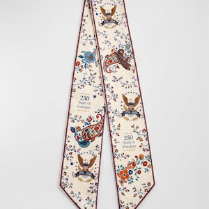 May include: Cream-coloured scarf with a repeating floral and paisley pattern. The scarf features the text "250 Years of Freedom" and the years "1776-2026", along with images of eagles. The edges are trimmed with a dark red border.