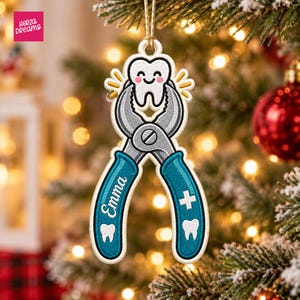 May include: A hanging ornament shaped like a pair of teal dental pliers holding a smiling white tooth. The pliers feature the name Emma in white script, a white medical cross, and small white tooth icons on the teal handles.