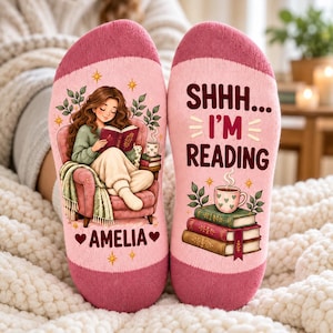 Op de afbeelding: Een paar lichtroze sokken met donkerroze boorden. De linkersok toont een illustratie van een lezend persoon in een stoel met de naam AMELIA. De rechtersok toont de tekst SHHH... I'M READING boven een stapel boeken.